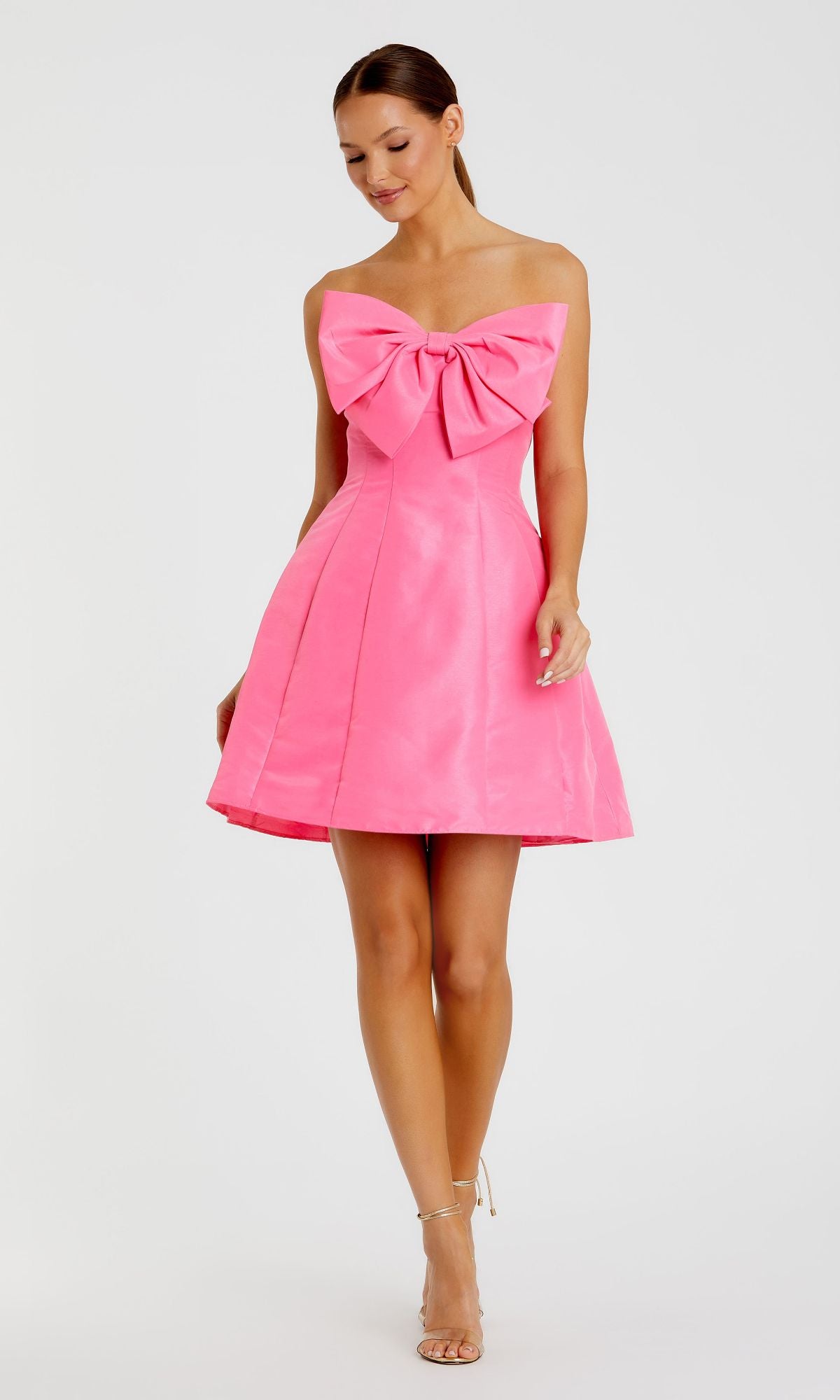 A woman in the Mac Duggal 49210 Strapless Short Bow Party Dress in bright pink, featuring a large chest bow, stands looking down and pairs it with nude heels.